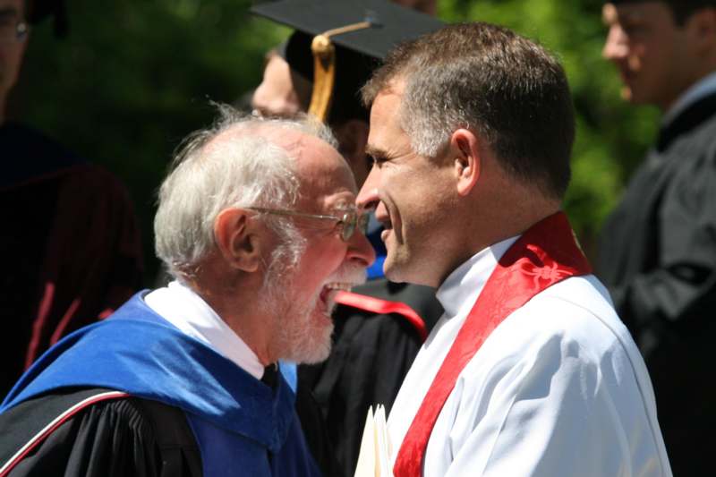 a man in a graduation gown laughing with another man in a cap