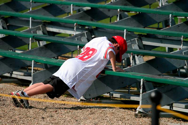 a boy in a white jersey and red hat leaning on a green bleachers