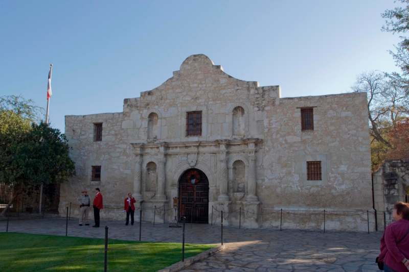 a stone building with a stone archway and people standing in front of it with Alamo Mission in San Antonio in the background