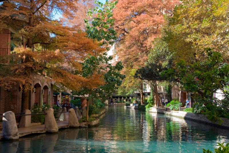 a river with trees and buildings with San Antonio River Walk in the background