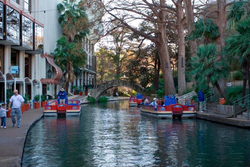 a group of boats on a canal with San Antonio River Walk in the background