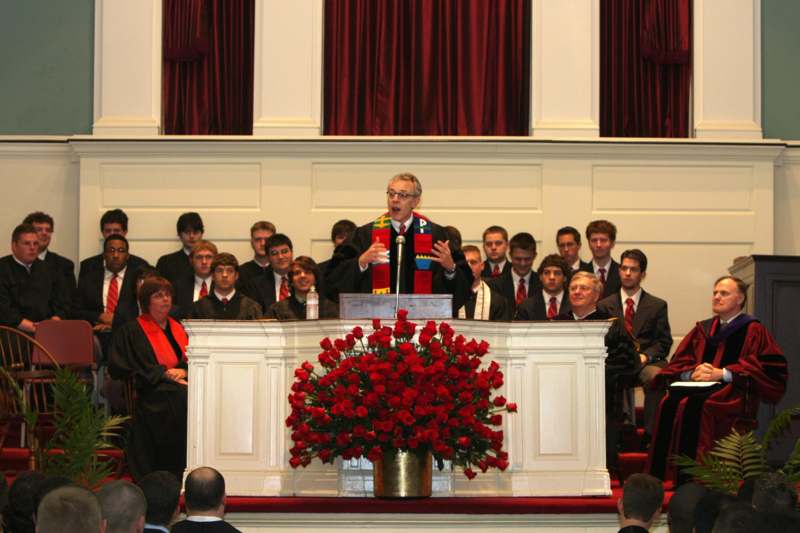 a man speaking into a microphone at a podium with a group of people in the background