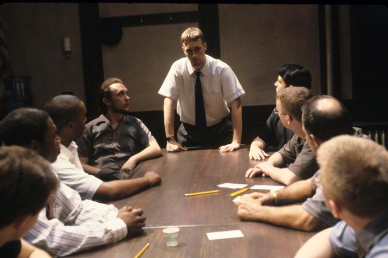 a man standing in front of a group of men sitting around a table