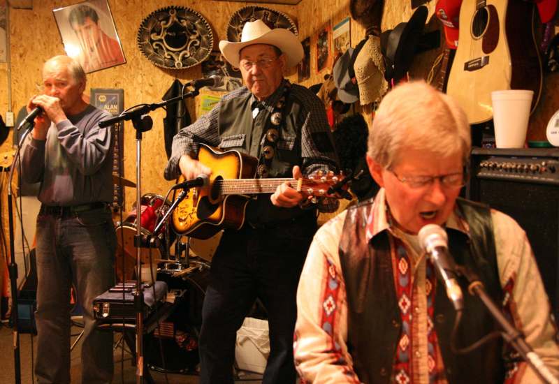a group of men singing in a room with a guitar and a man in a cowboy hat