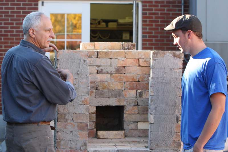 a man looking at another man standing next to a brick oven