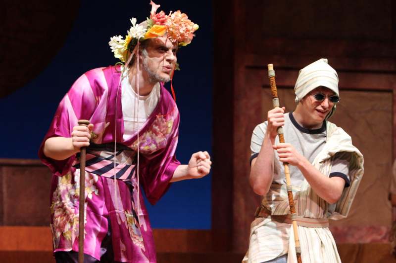 a man in a flower crown holding a stick and a man in a dress