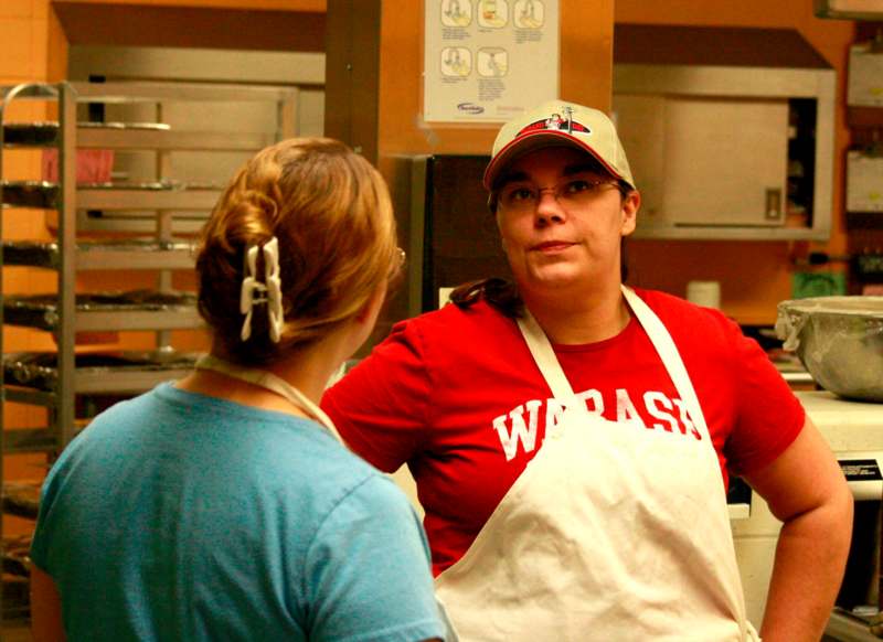 a woman in a white apron talking to a woman in a kitchen