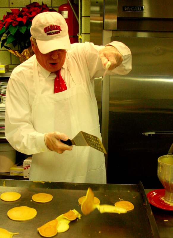 a man in a white apron and red tie cooking pancakes