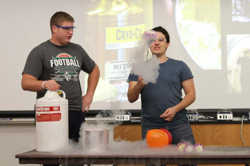 a man standing next to another man standing next to a white cylinder with smoke