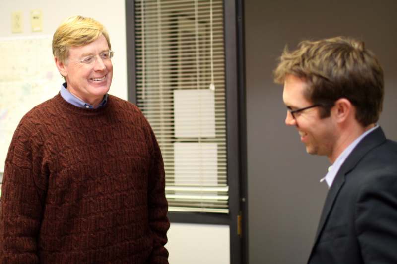a man in a brown sweater and glasses talking to another man