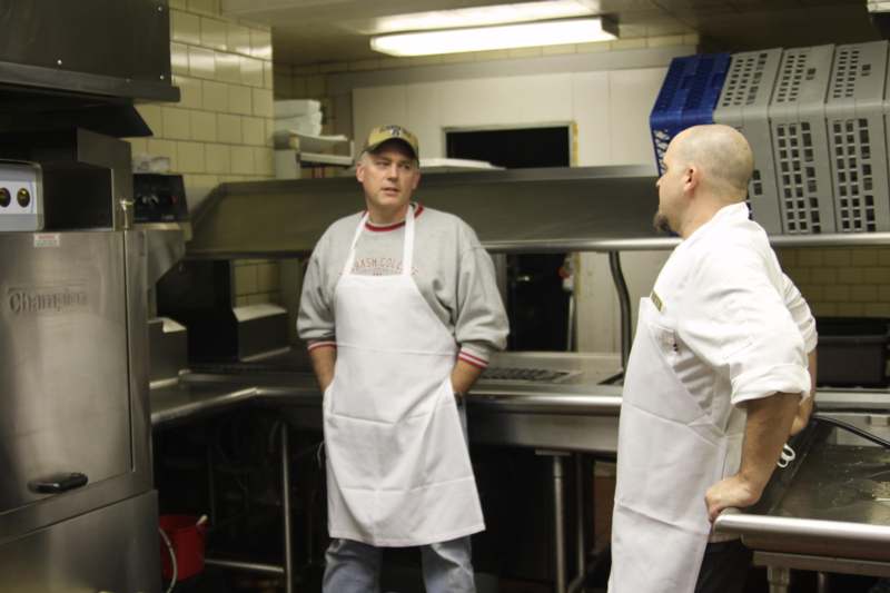 a man in a white apron standing next to a man in a kitchen