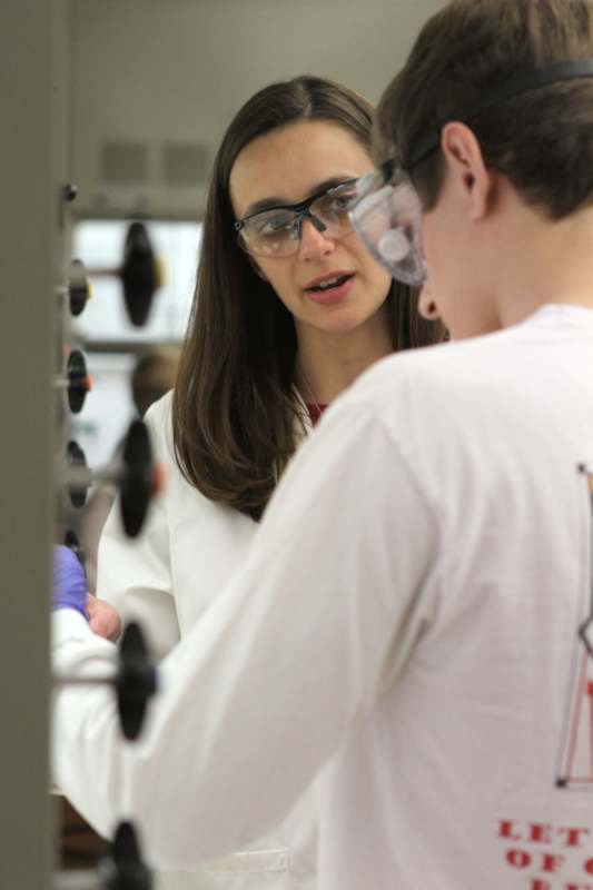 a woman in white lab coat and goggles talking to a man in a lab