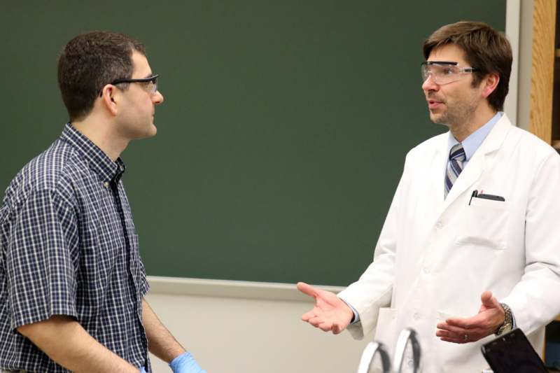 a man in a white coat and goggles talking to another man