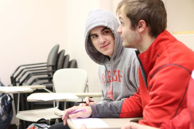 a man in a hoodie sitting at a desk with another man in a hoodie