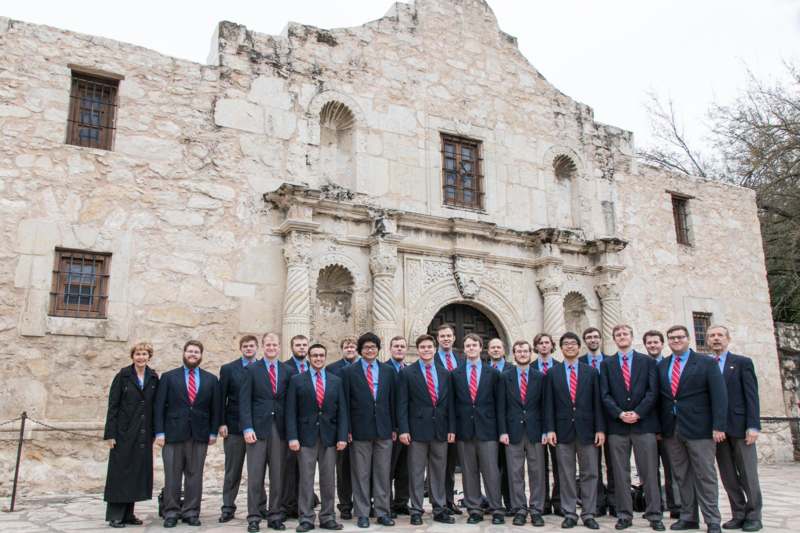 a group of men in suits standing in front of a stone building with Alamo Mission in San Antonio in the background