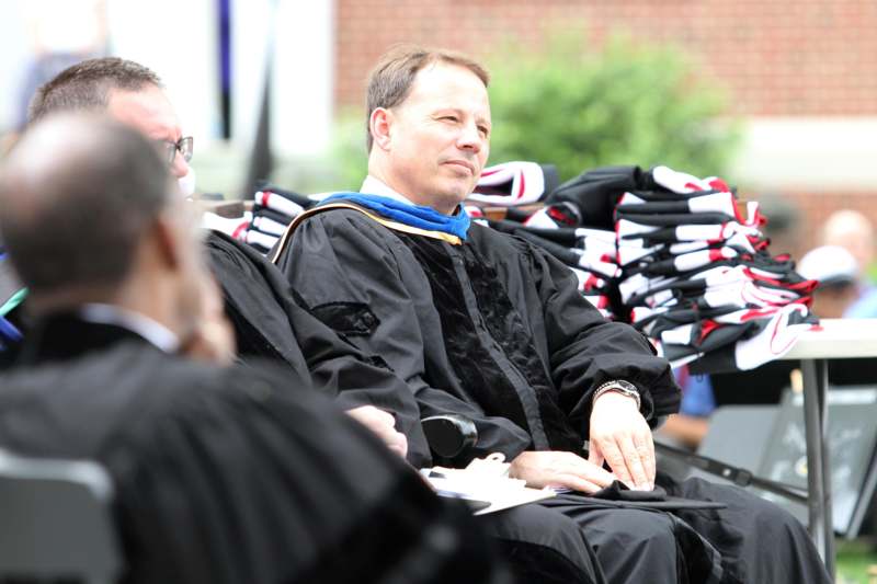 a man in a black robe sitting in a chair with stacks of stacks of graduation caps