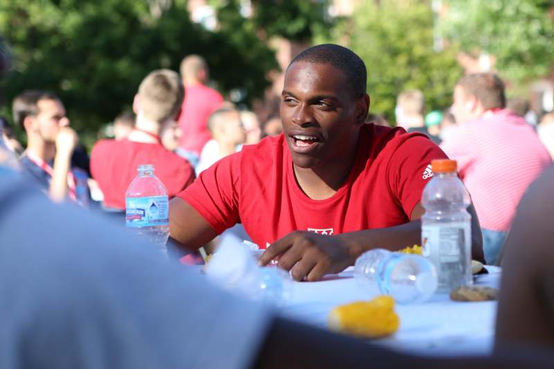 a man in red shirt sitting at a table with water bottles and a group of people