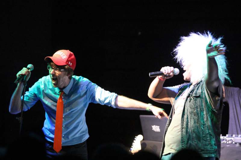 a man singing into a microphone next to a man with a wig on