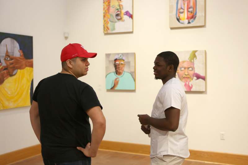 a man in a red hat talking to another man in a room with paintings on the wall