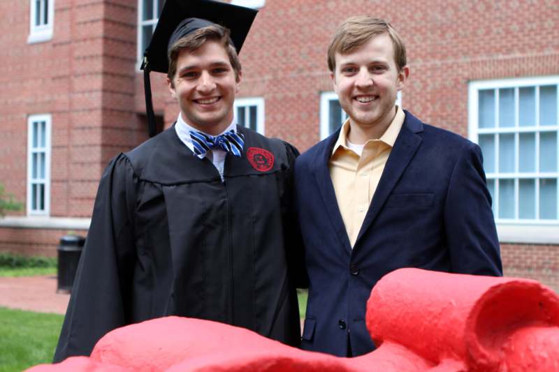 two men in graduation gowns and cap standing next to a red statue