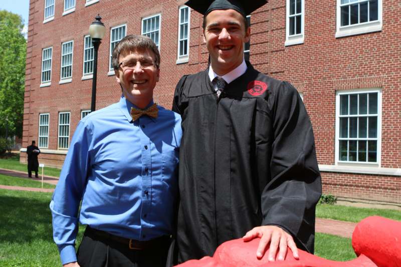 a man in a graduation gown and cap standing next to a man in a blue shirt
