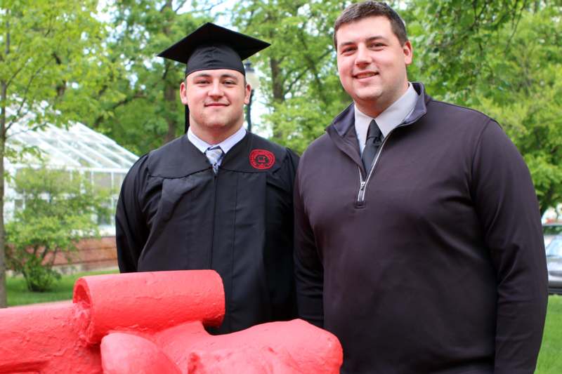two men in graduation gowns and cap standing next to a red statue
