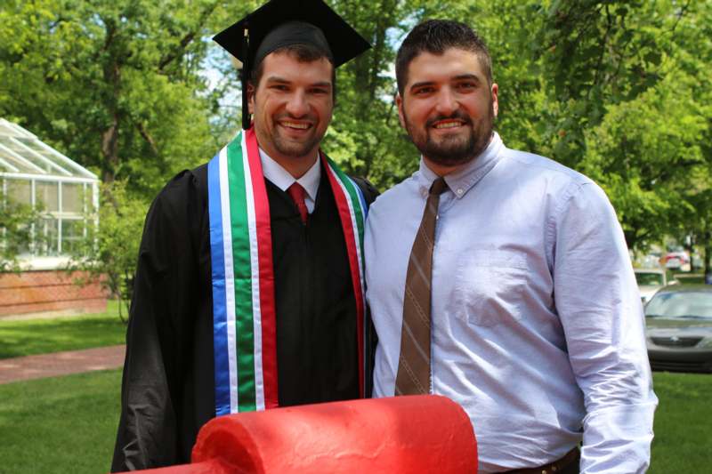 a man in a graduation gown and cap standing next to another man in a park