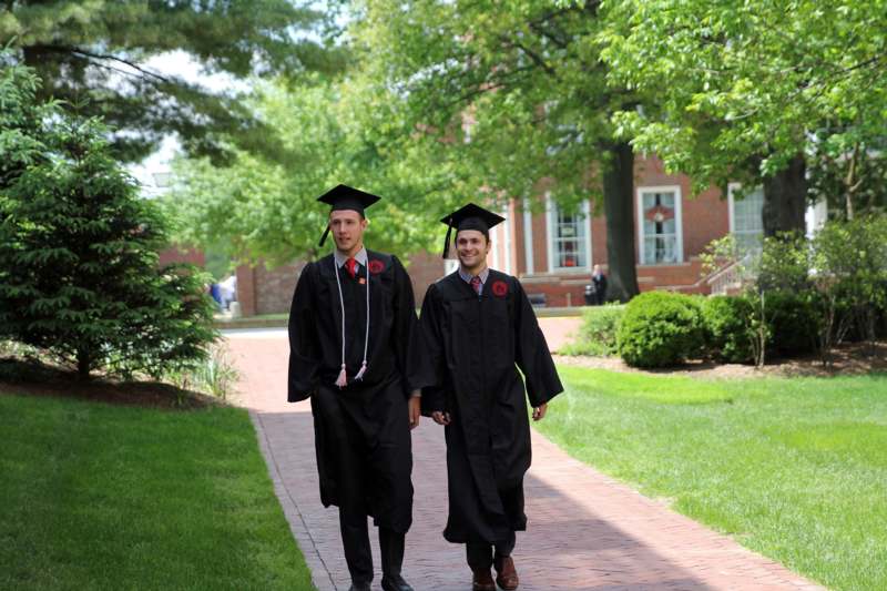 two men in graduation gowns and caps walking down a brick path