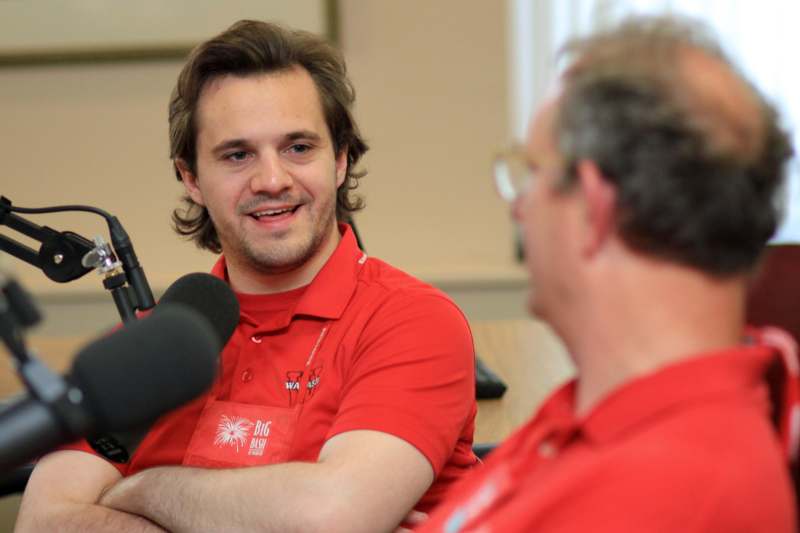 a man in a red shirt talking to another man in a room with microphones