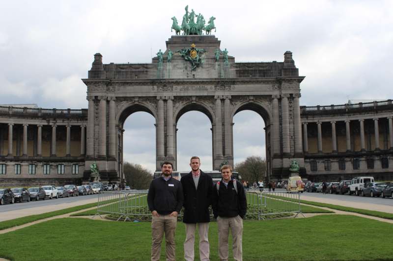 a group of men standing in front of a large stone structure with Cinquantenaire in the background