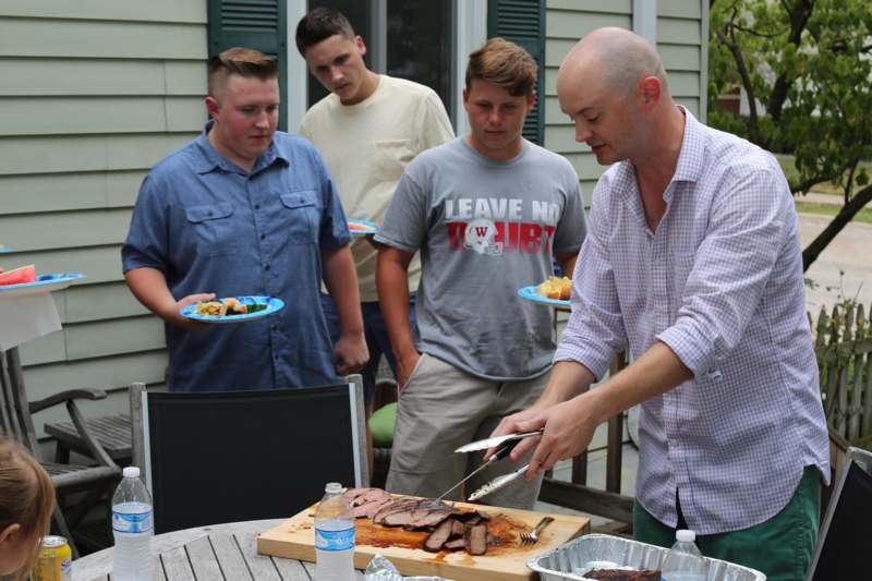 a group of men standing around a table with food and a man holding tongs