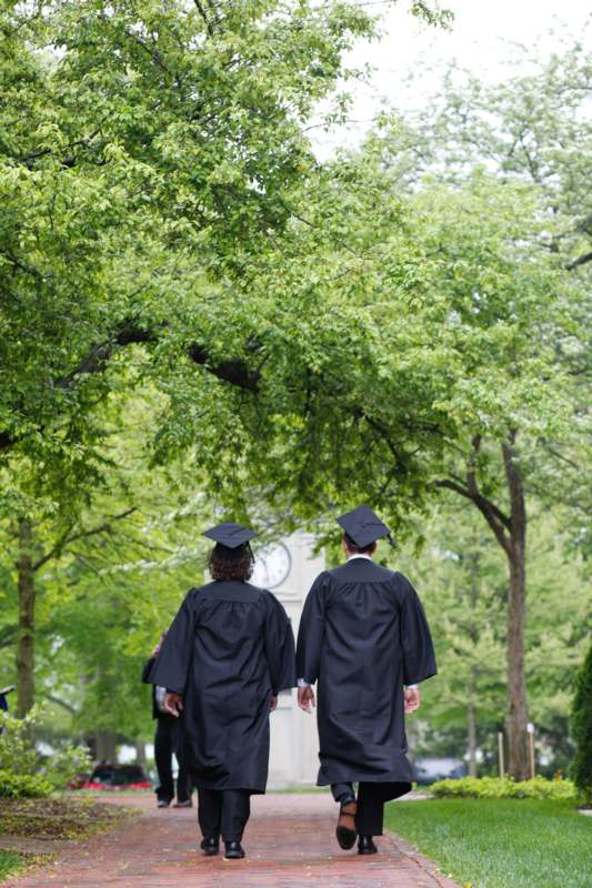 two people in graduation gowns and caps walking down a sidewalk