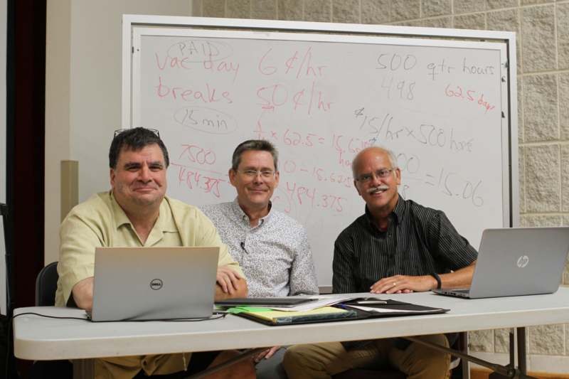 a group of men sitting at a table with laptops and a whiteboard
