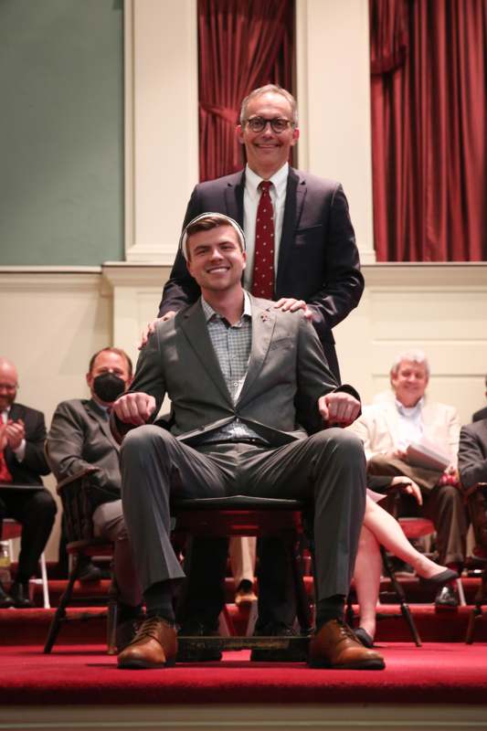 a man sitting on a chair with another man in a suit
