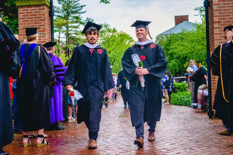 two men in graduation gowns and caps walking down a brick path