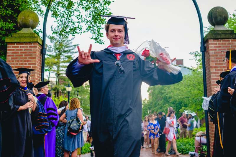 a man in a graduation gown holding flowers and giving a hand gesture