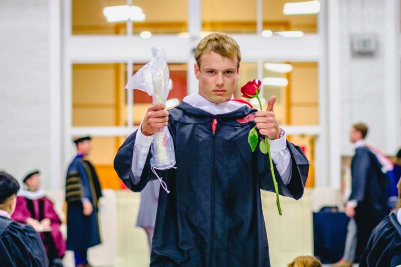 a man in a graduation gown holding a rose and giving a thumbs up