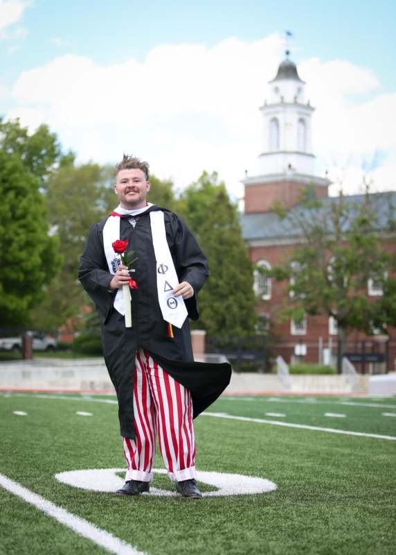a man in a graduation gown holding a rose and standing on a football field