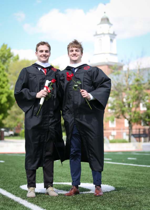 two men wearing graduation gowns and holding roses