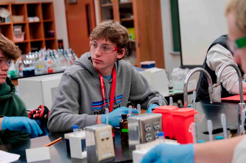 a man wearing safety glasses and gloves sitting at a table with other people in the background