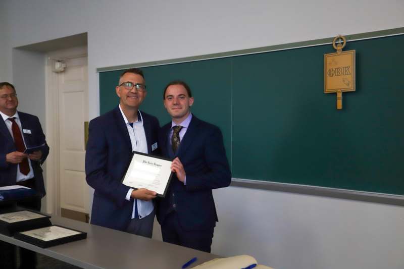 two men in suits holding a certificate in front of a chalkboard