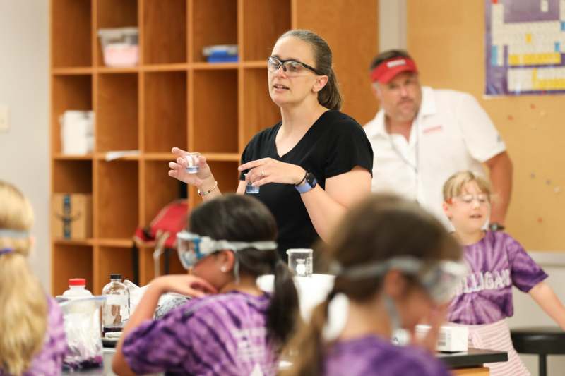 a woman in a black shirt and glasses holding a small glass with a child in a classroom