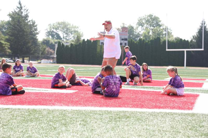 a man in a white shirt and tan shorts talking to a group of boys sitting on a field