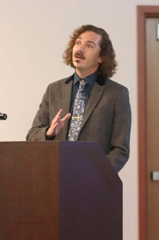 a man with long hair and mustache standing at a podium
