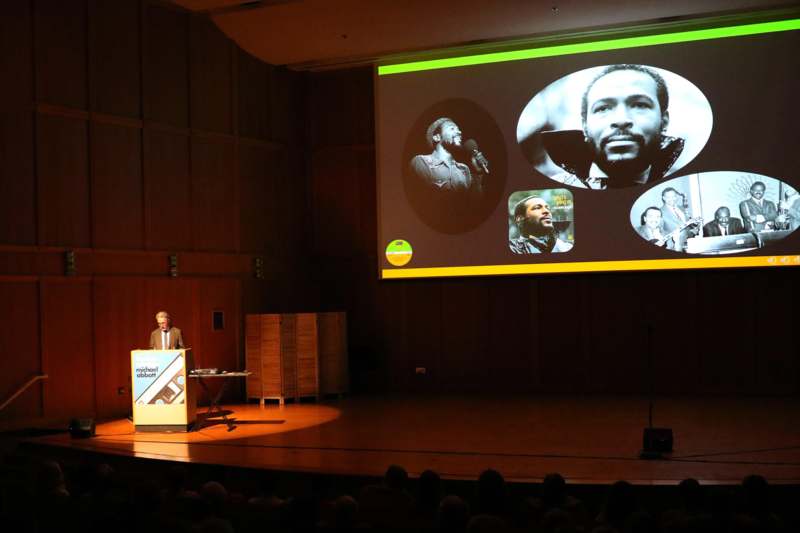 a man standing at a podium with a large screen on the wall