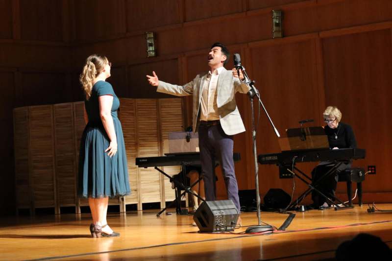 a man singing on a stage with a woman and a piano