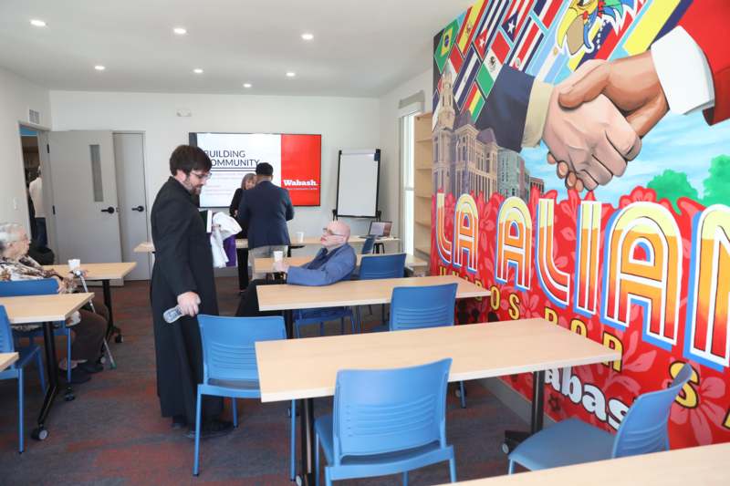 a man in a black robe standing in a room with chairs and a poster on the wall