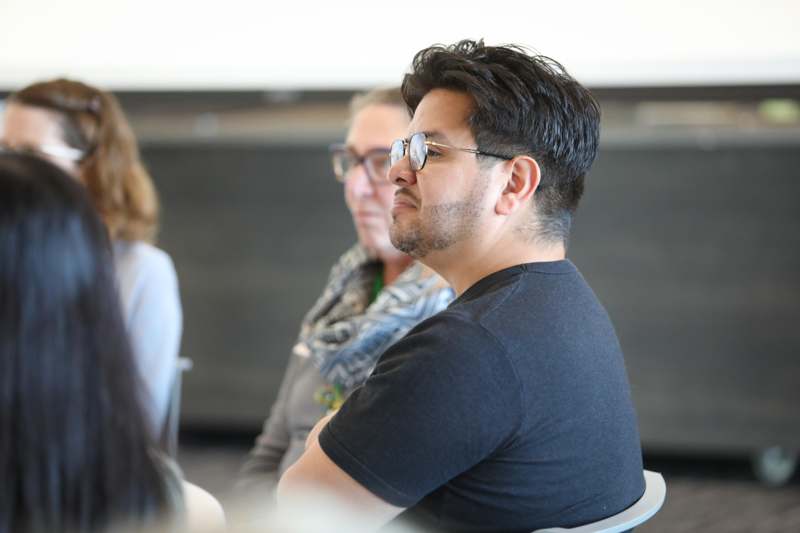 a man wearing glasses and a black shirt sitting in a room with other people