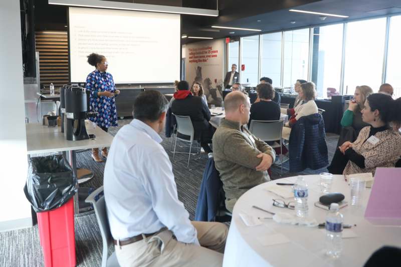 a woman standing in front of a group of people sitting around a table