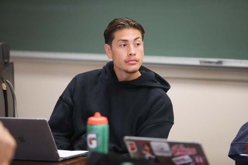 a man sitting at a desk with a laptop and a green chalkboard